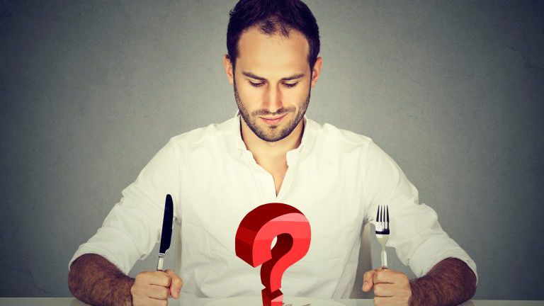 Mann mit Gabel und Messer sitzt am Tisch und blickt auf Teller mit großer roter Frage