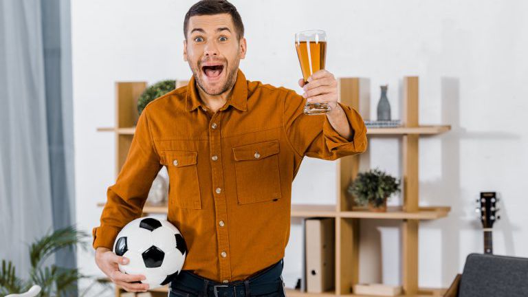 schöner Mann in modernem Wohnzimmer schreit, während er Fußball und Bier in der Hand hält