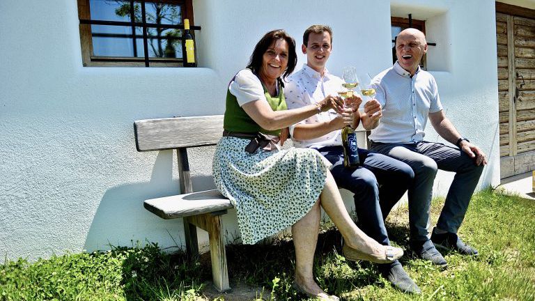 Eine Frau und zwei Männer auf einer Bank mit Weingläsern, zuprosten
